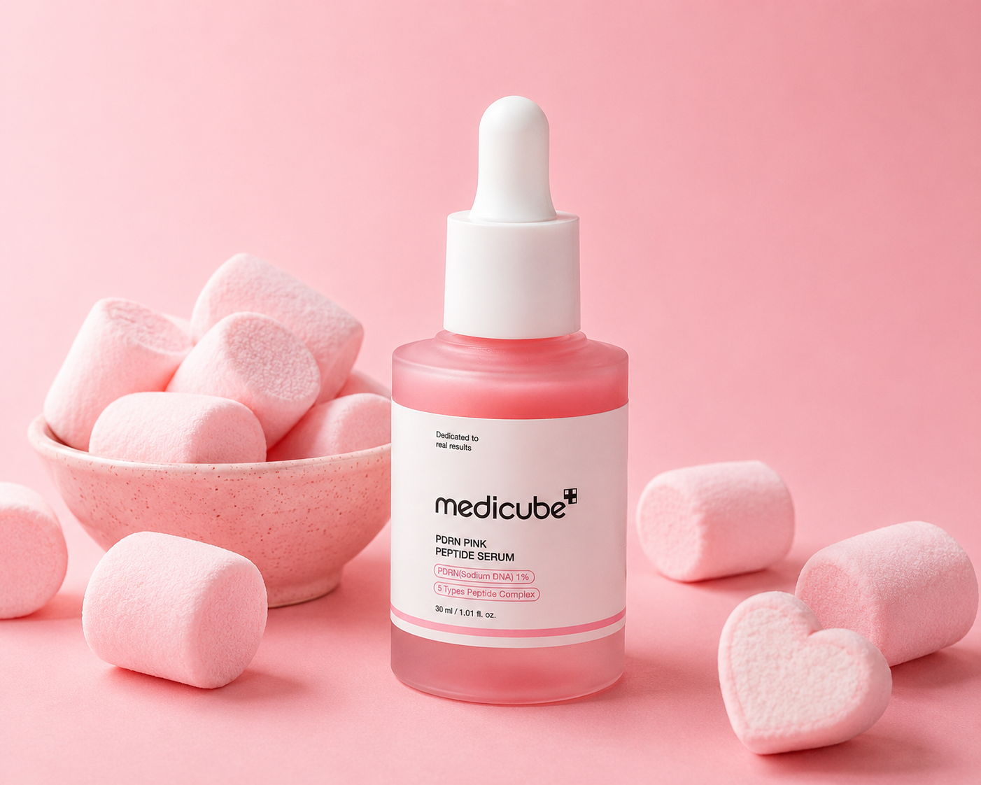Medicube PDRN Pink Peptide Serum 30ml glass dropper bottle with a soft pink translucent formula, displayed upright among scattered pink and white marshmallows and a heart-shaped marshmallow on a pastel pink background