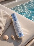 VT Cica Reedle Shot 100 microneedling serum in a sleek silver airless pump bottle, 50mL, resting beside a folded white towel and two seashells on a poolside surface with sparkling blue water in the background